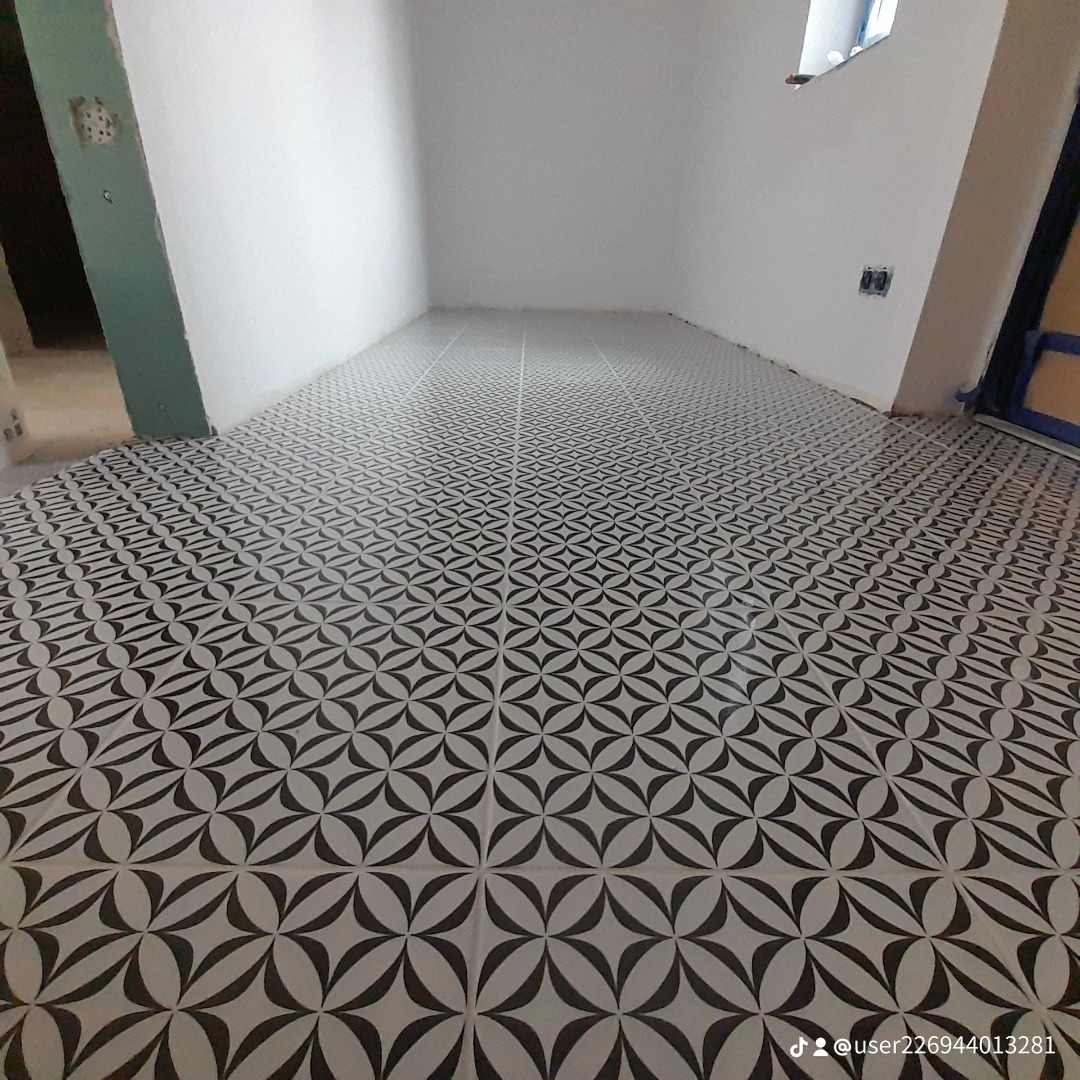 Ułożona podłoga z czarno-białych, wzorzystych płytek w pustym pomieszczeniu z białymi ścianami, widoczny fragment okna i drzwi zabezpieczonych taśmą malarską.