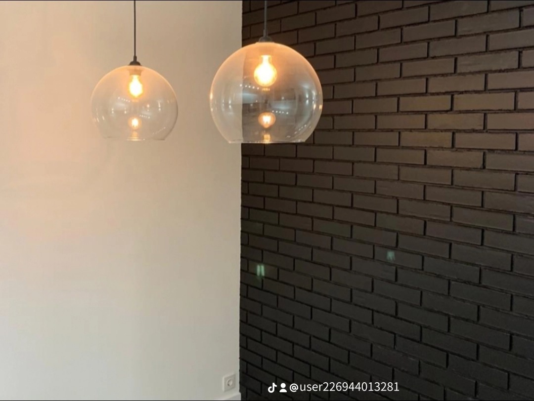 Nowoczesna ściana z czarnej cegły w pomieszczeniu z dwiema szklanymi lampami wiszącymi i białą ścianą, widoczny fragment gniazdka elektrycznego.