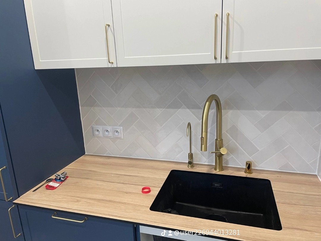 Nowoczesna kuchnia z granatowymi szafkami dolnymi i białymi górnymi, złotą armaturą i czarnym zlewem. Na blacie leży czerwona uszczelka i wizytówka.