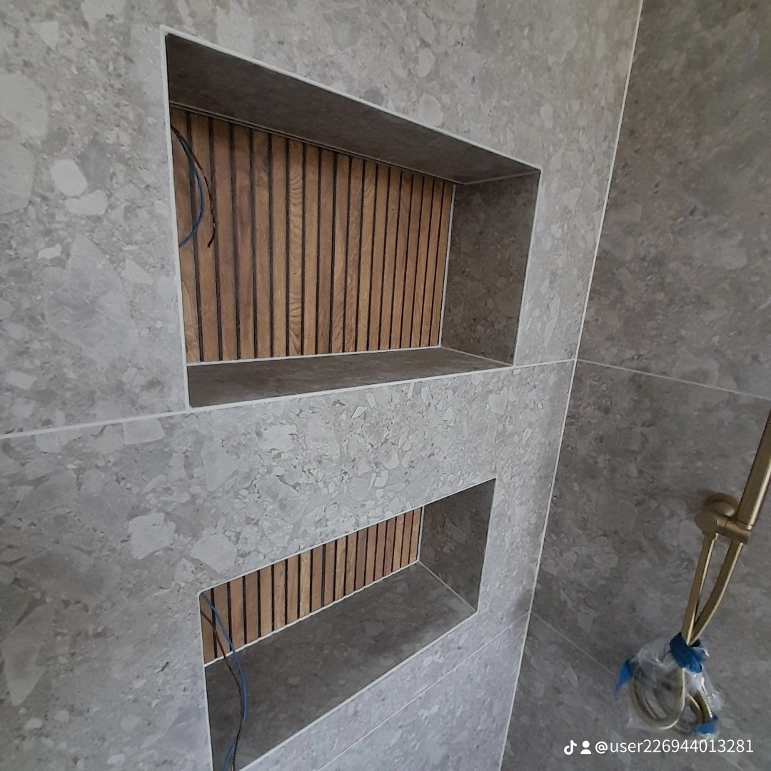 Dwa wnęki w ścianie łazienki wyłożonej szarymi płytkami imitującymi kamień, wykończone drewnianymi panelami pionowymi; widoczny fragment słuchawki prysznicowej w kolorze złotym.