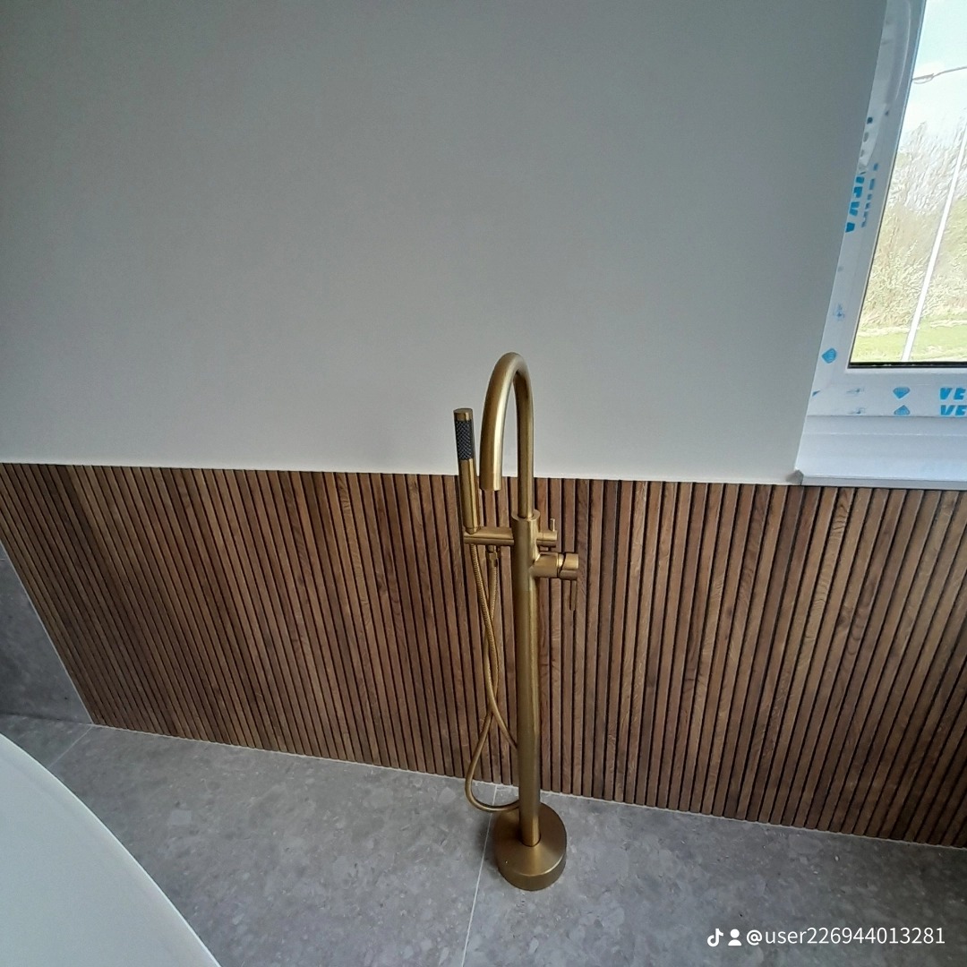Wolnostojąca, złota bateria wannowa z prysznicem na tle drewnianej boazerii i szarej płytki podłogowej w jasnej łazience, widok z góry na baterię i fragment wanny.