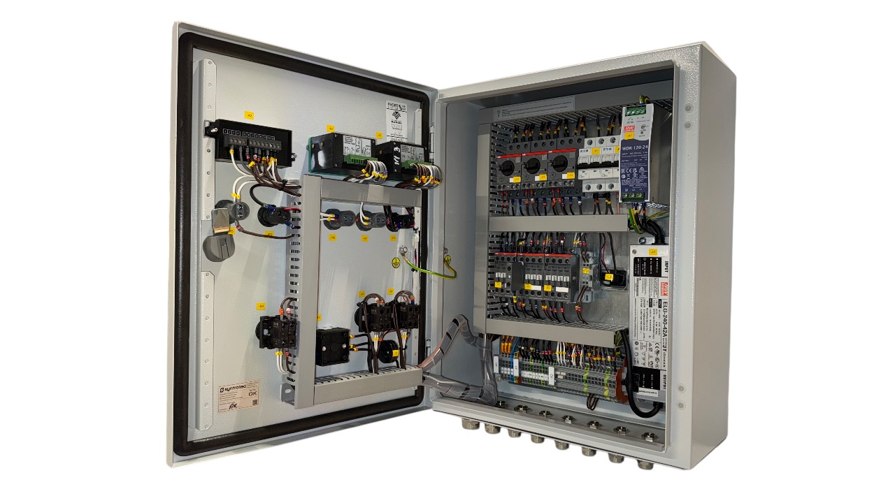 Otwarta szafa sterownicza z widocznymi elementami elektrycznymi, takimi jak bezpieczniki, przekaźniki i okablowanie. Widoczne oznaczenia na elementach.
