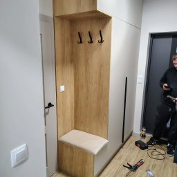Funkcjonalna szafa w przedpokoju z siedziskiem i wieszakami, wykończona w drewnie i bieli. Widoczne narzędzia montażowe na podłodze.