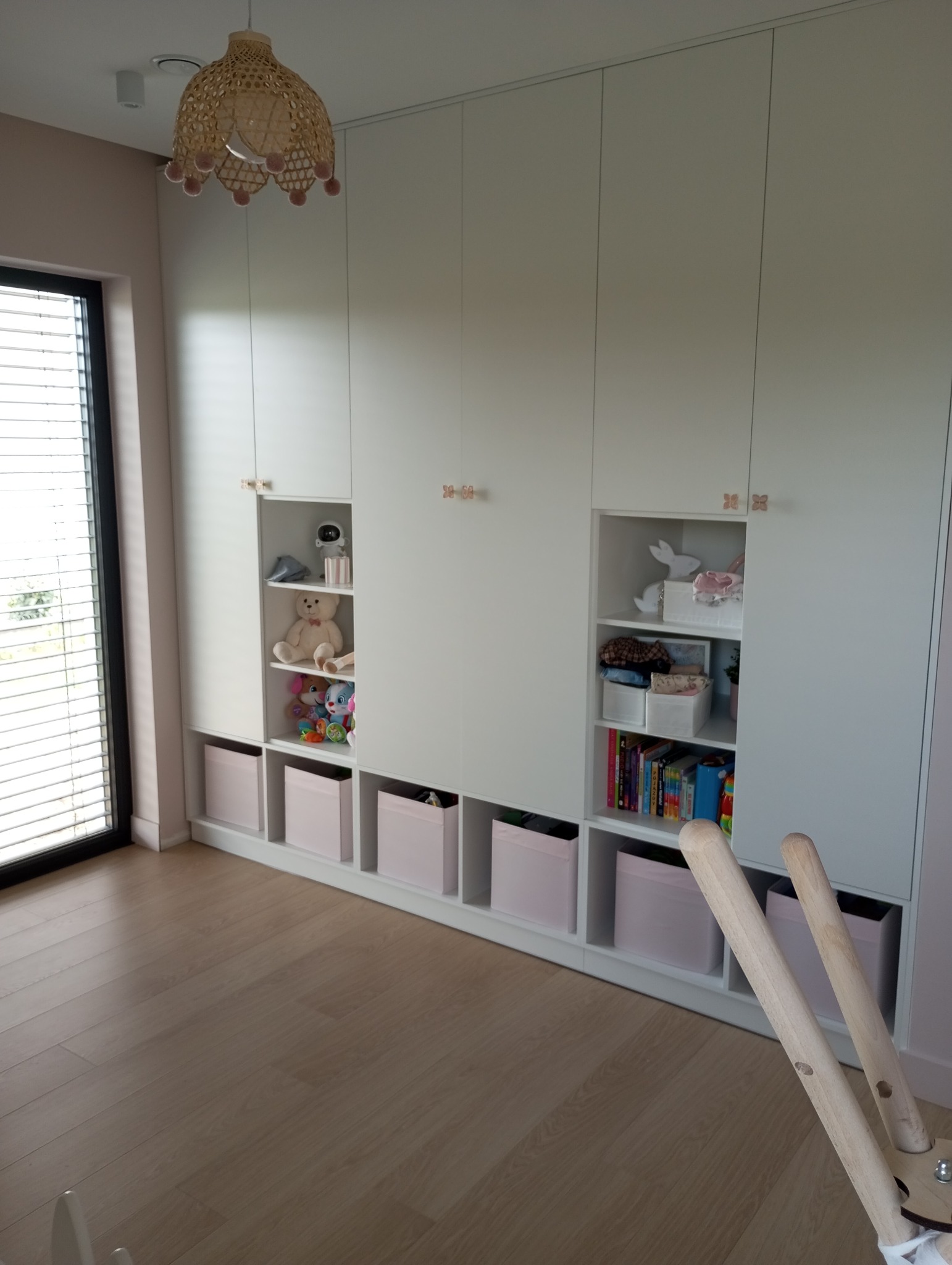 Biała szafa wnękowa z otwartymi półkami, na których znajdują się zabawki i książki. Pod półkami widoczne różowe pojemniki. Całość w pokoju dziecięcym.
