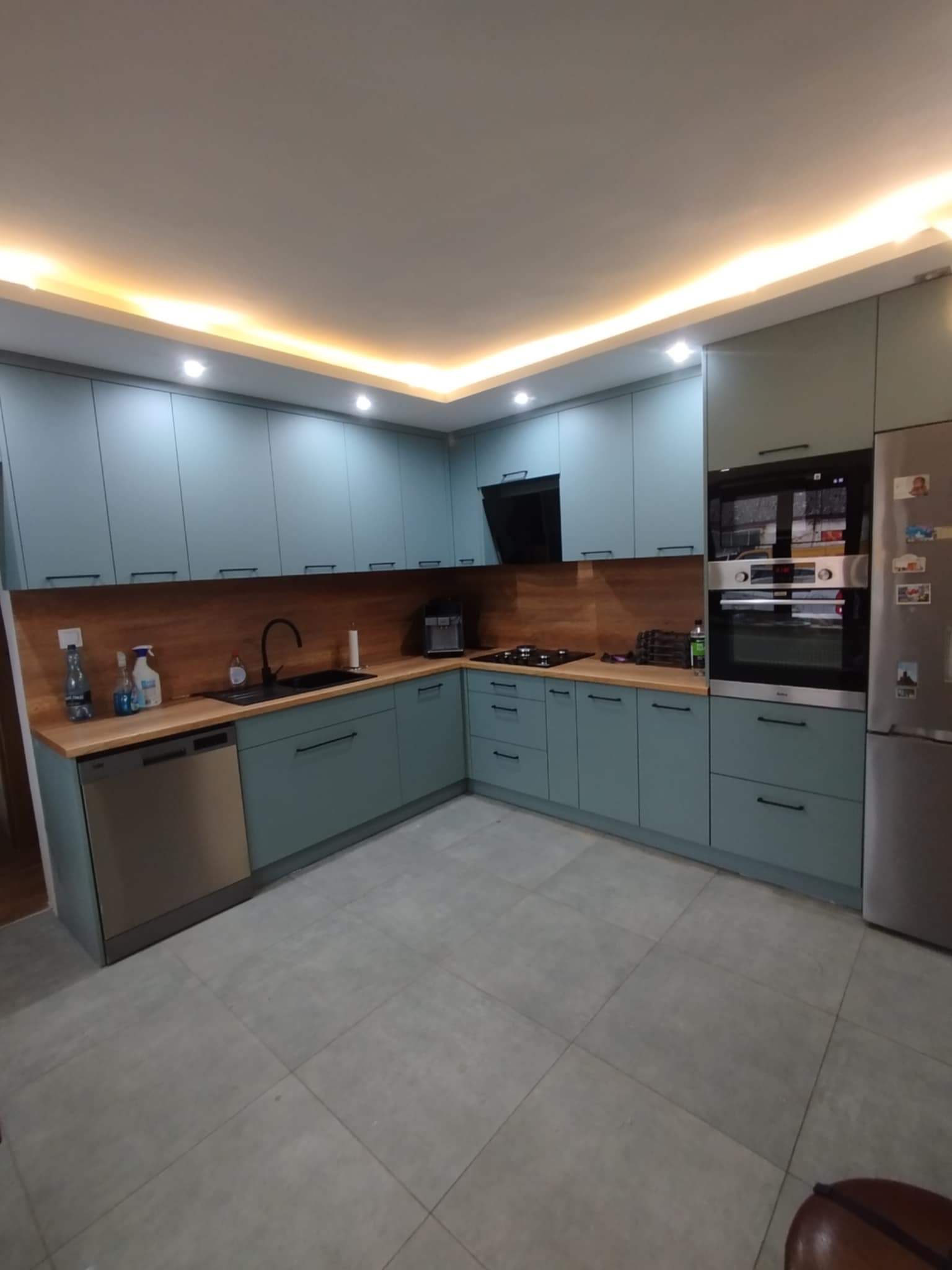 Turkusowa kuchnia z drewnianym blatem i oświetleniem LED podszafkowym, widok na piekarnik i lodówkę. Nowoczesny design z czarną armaturą.