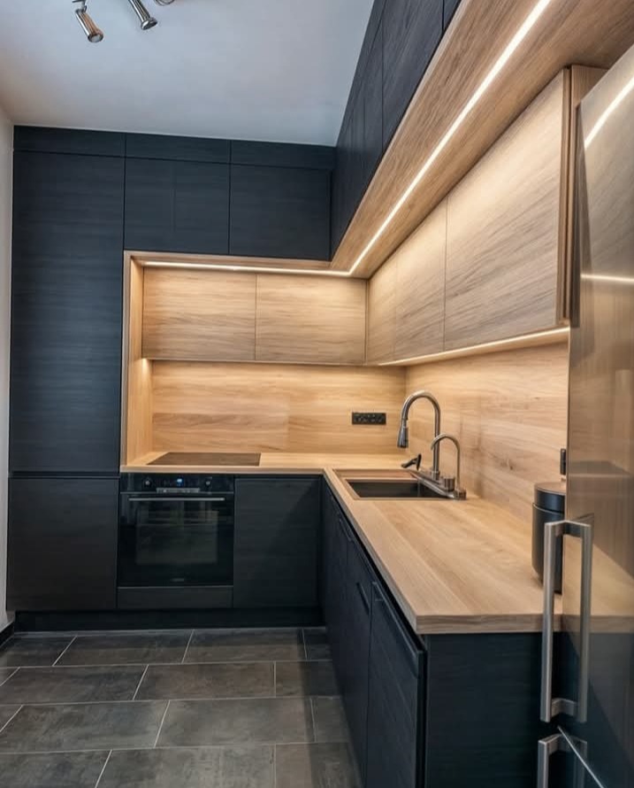 Nowoczesna kuchnia z ciemnymi szafkami i drewnianymi blatami, podświetlona listwami LED. Minimalistyczny design z czarną płytą grzewczą i zlewozmywakiem narożnym.
