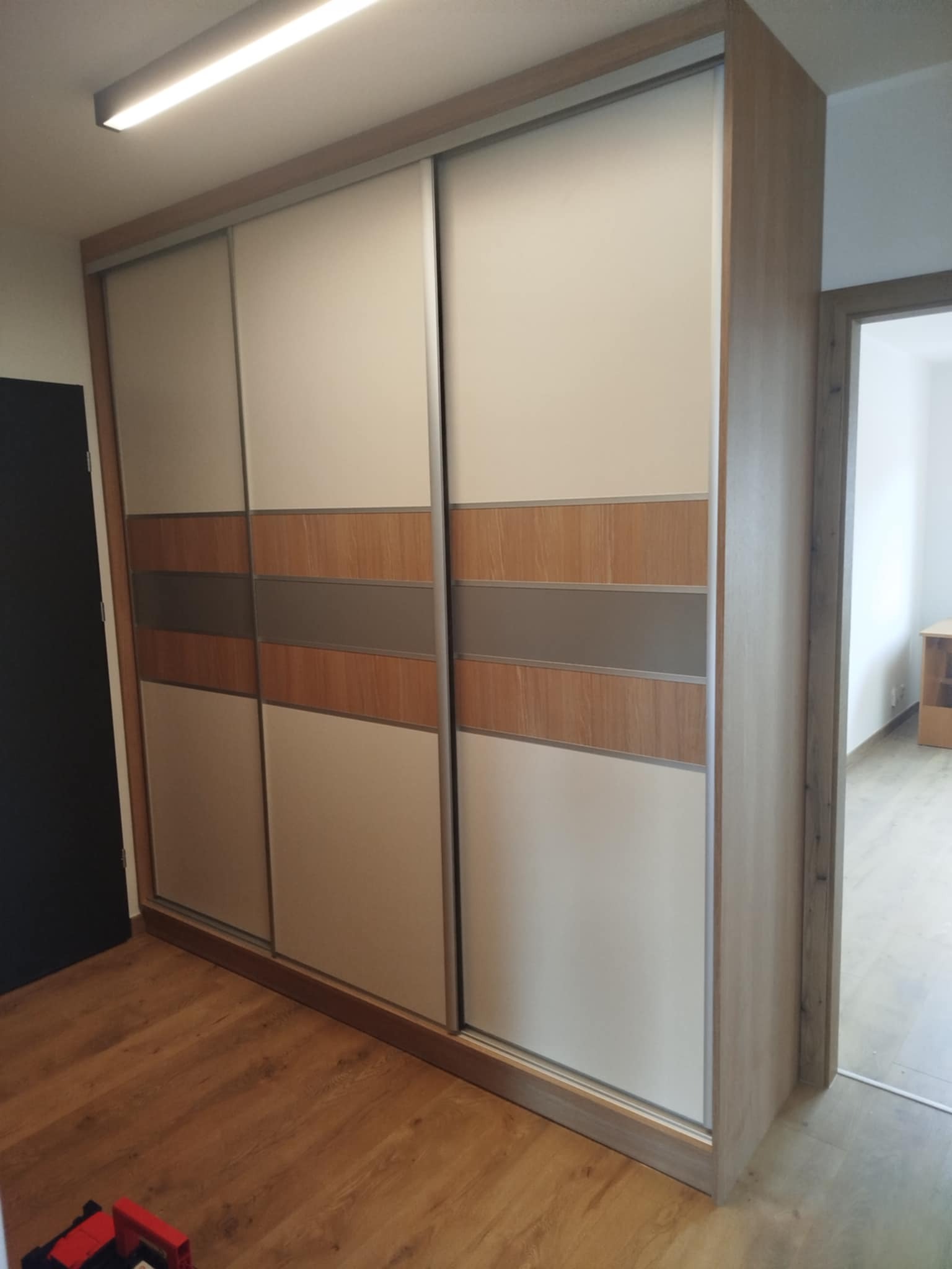 Jasna szafa wnękowa z przesuwnymi drzwiami, z poziomymi pasami w odcieniach drewna i szarości, na tle drewnianej podłogi i częściowo widocznych drzwi.