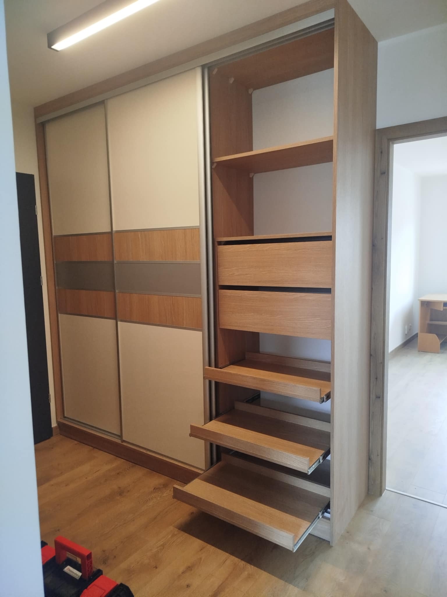 Jasna szafa wnękowa z przesuwnymi drzwiami i wysuwanymi półkami, widoczna z boku. Na podłodze czerwona skrzynka narzędziowa. W tle widok na puste pomieszczenie.