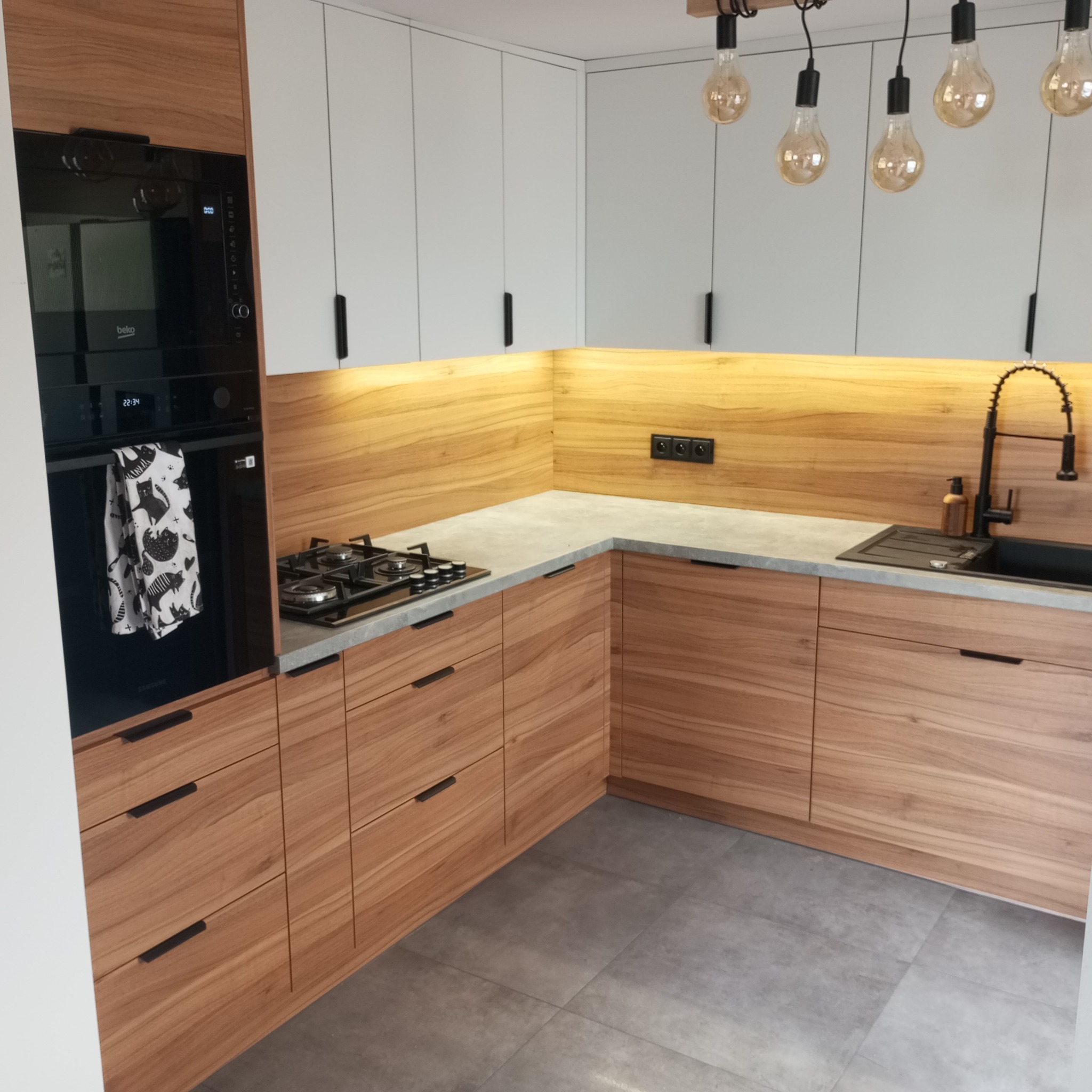 Nowoczesna kuchnia na wymiar z drewnianymi frontami i białymi szafkami wiszącymi, oświetlona lampami Edison. Na blacie płyta gazowa i czarny zlew.