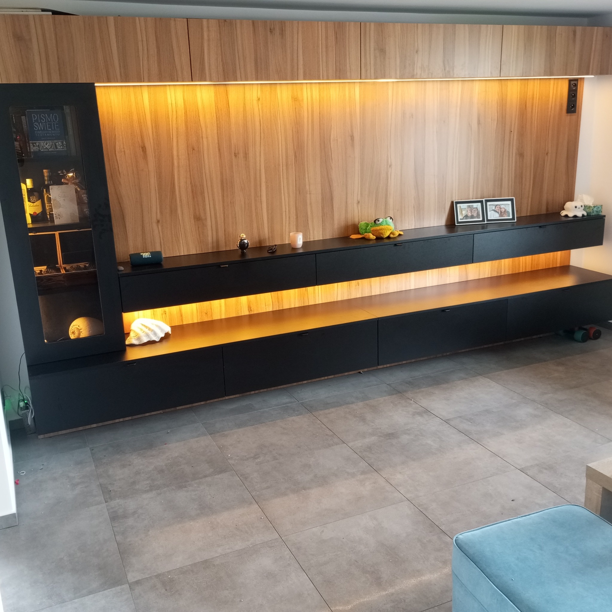 Nowoczesna zabudowa meblowa w salonie: czarne szafki z drewnianym panelem i podświetleniem LED, z elementami dekoracyjnymi, w tym przeszklona witryna z alkoholem.