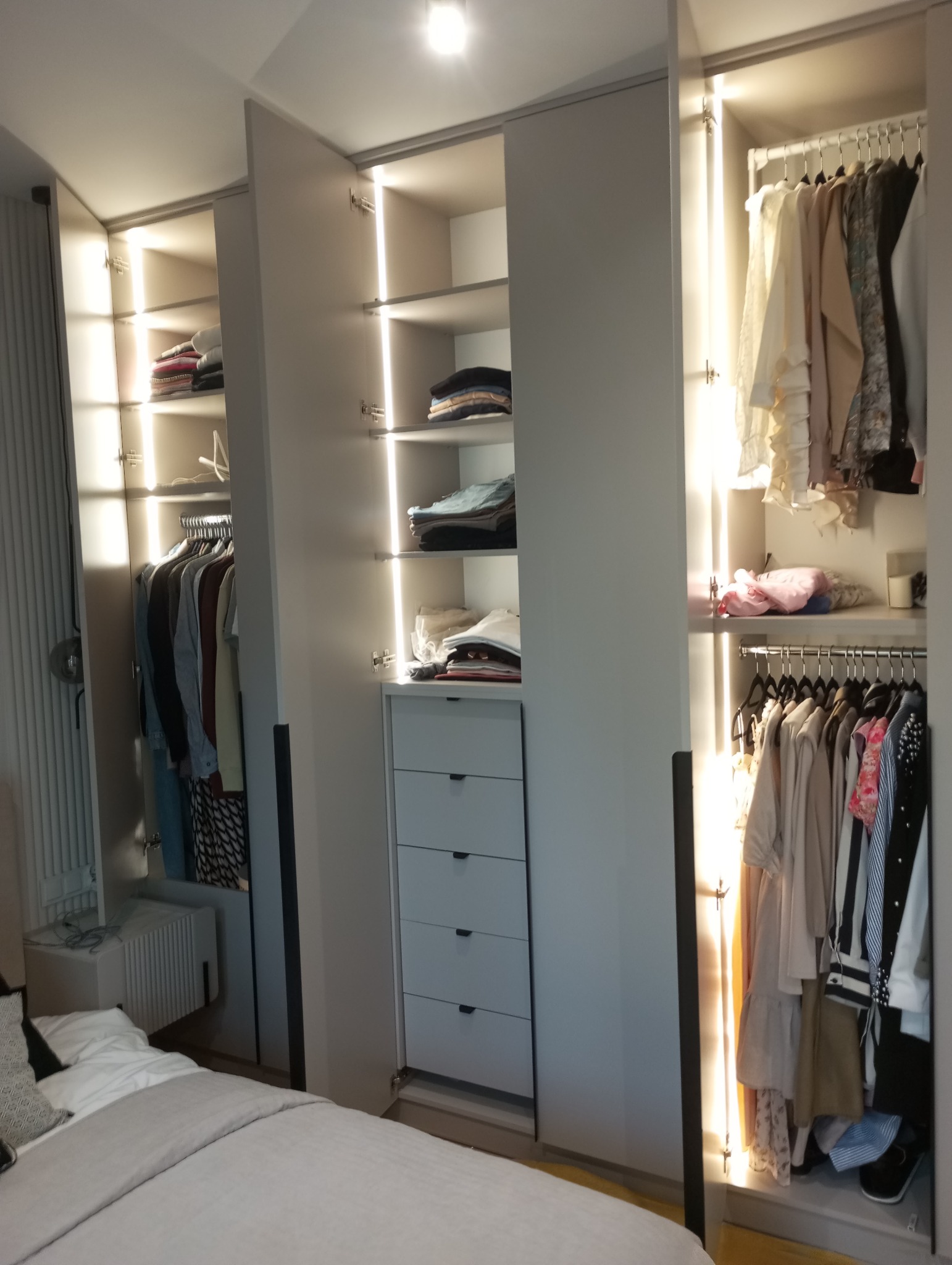 Jasna szafa wnękowa z oświetleniem LED, półkami i drążkami na ubrania, widoczna zawartość szafy oraz fragment łóżka w sypialni. Minimalistyczny design.