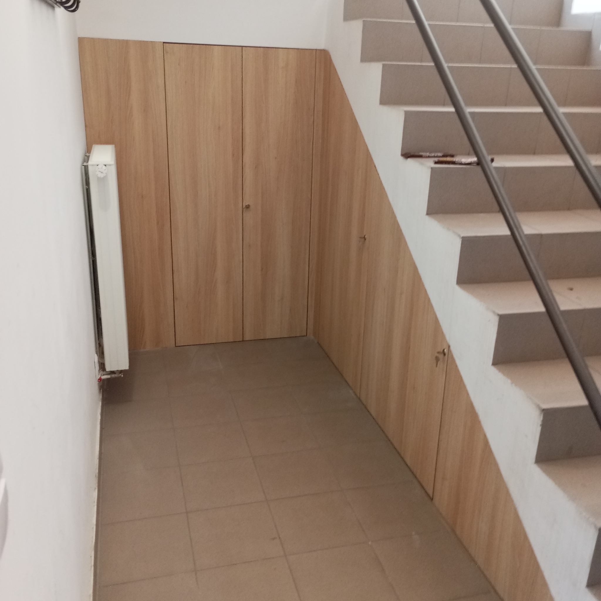 Drewniane szafki wnękowe pod schodami, z widocznym grzejnikiem i fragmentem metalowej poręczy. Jasna, kafelkowana podłoga. Ujęcie z dołu.