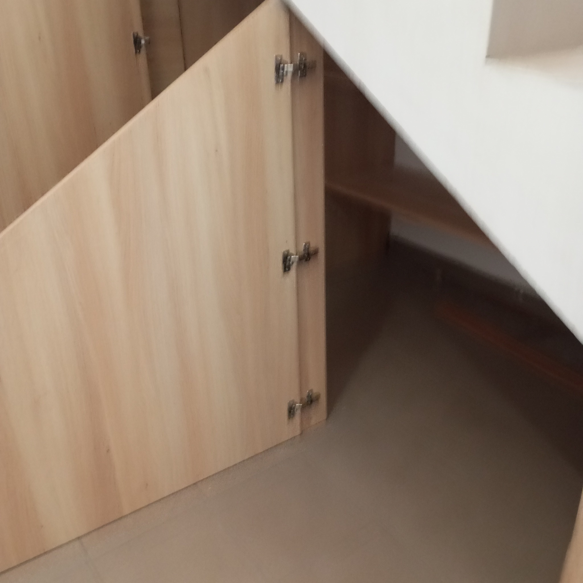 Szafka pod schodami z jasnego drewna, widoczne zawiasy, ukośny kształt dopasowany do przestrzeni. Funkcjonalne zagospodarowanie przestrzeni.