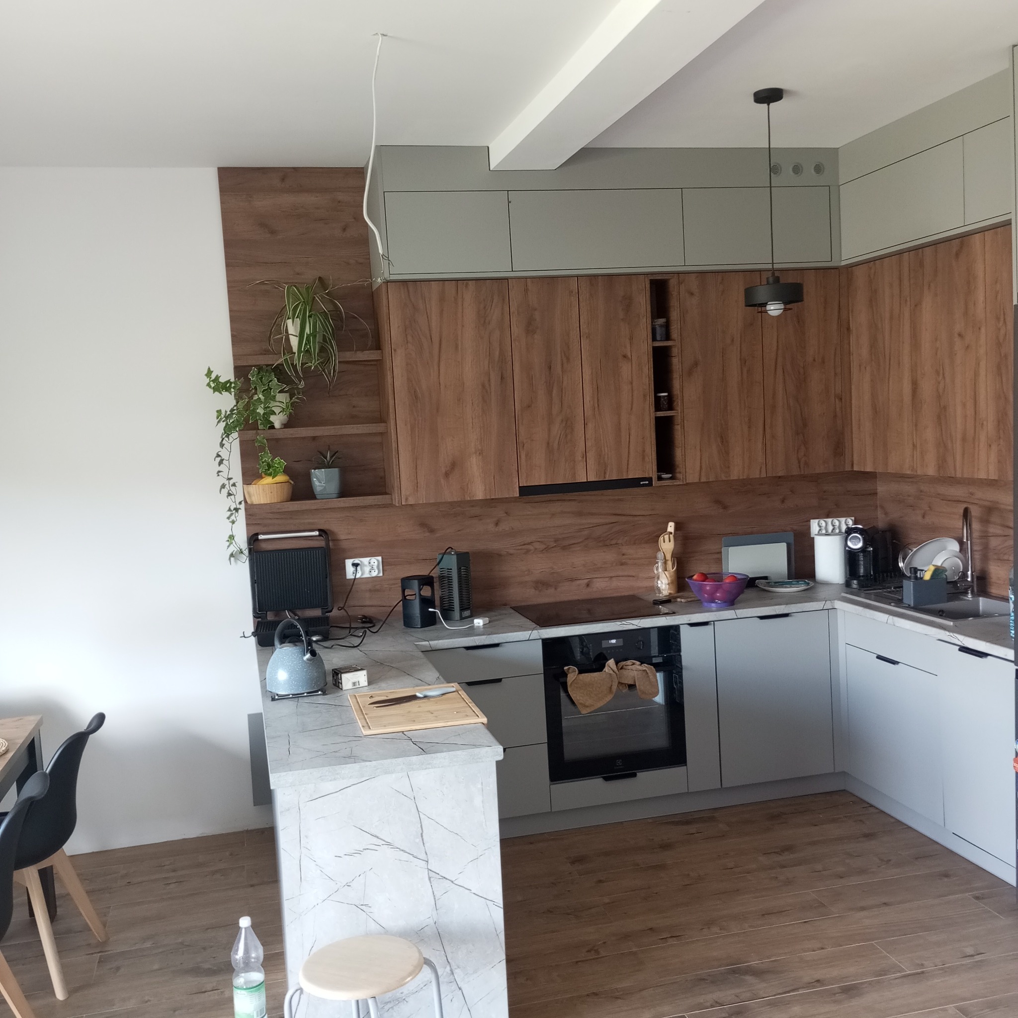 Nowoczesna kuchnia z drewnianymi frontami szafek i szarymi szafkami górnymi. Na blacie marmurowym deska do krojenia i sprzęt AGD. Wiszące rośliny dodają wnętrzu świeżości.