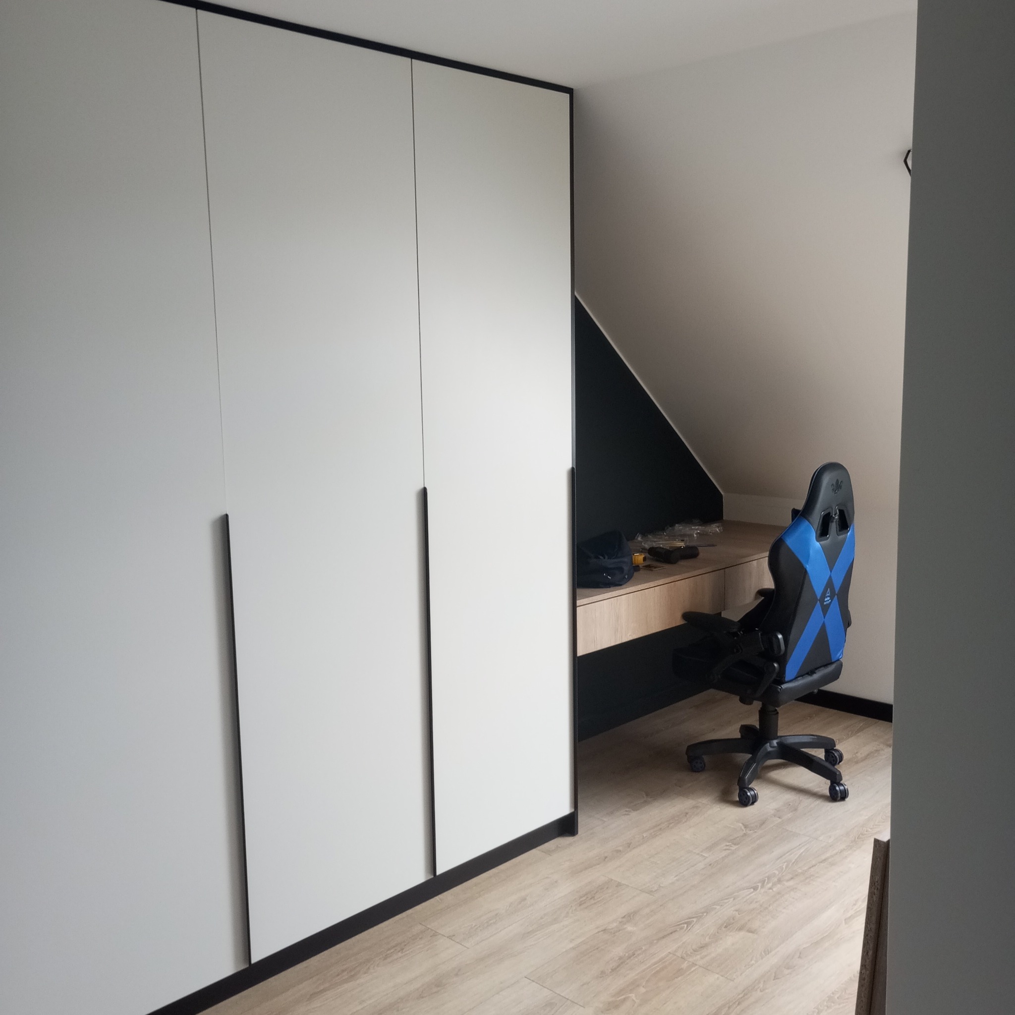 Biała szafa z czarnymi obramowaniami i długimi uchwytami w pokoju na poddaszu, obok biurko i krzesło gamingowe. Minimalistyczny design.