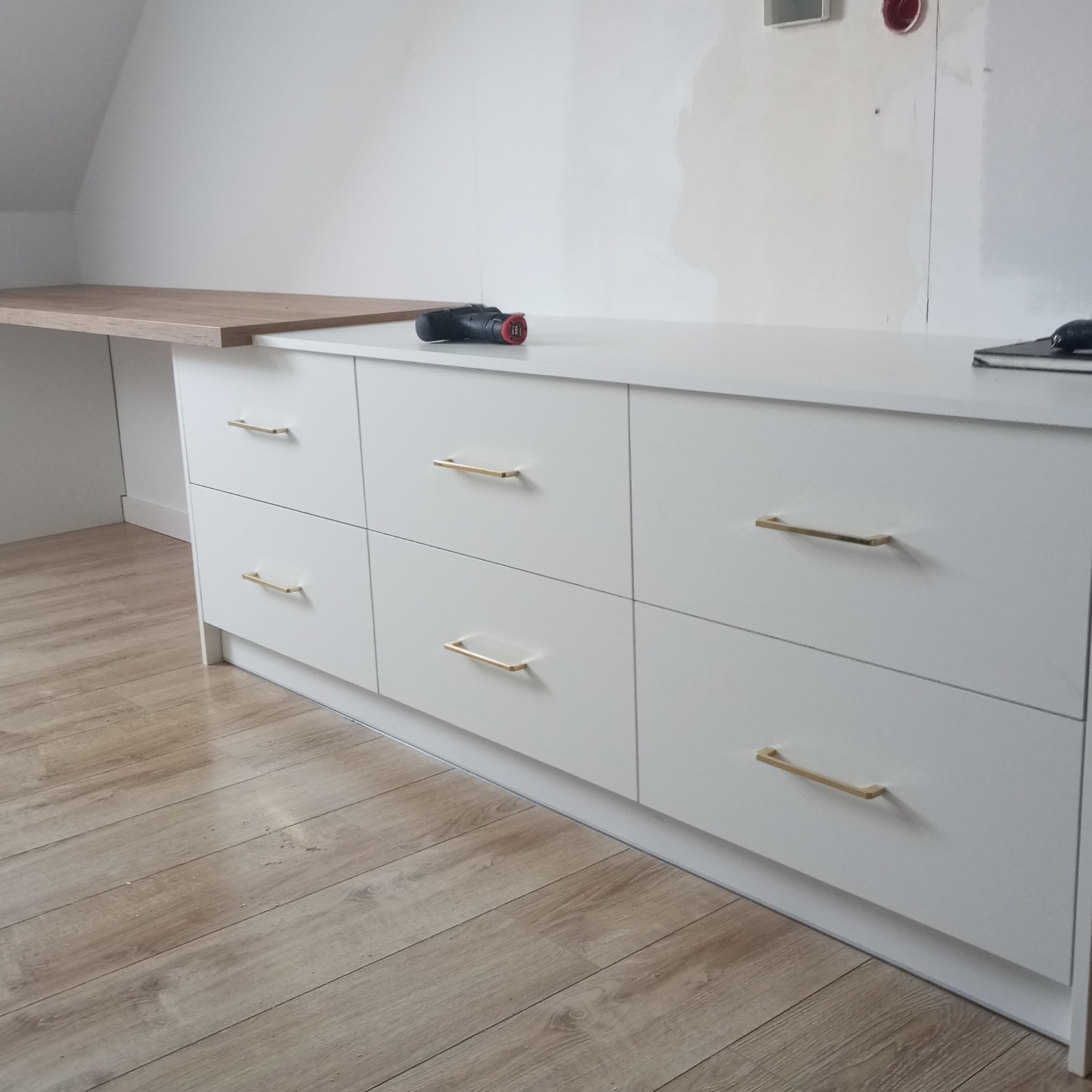 Biała komoda z sześcioma szufladami i złotymi uchwytami, uzupełniona drewnianym blatem i narzędziami, na tle białej ściany i podłogi z paneli.