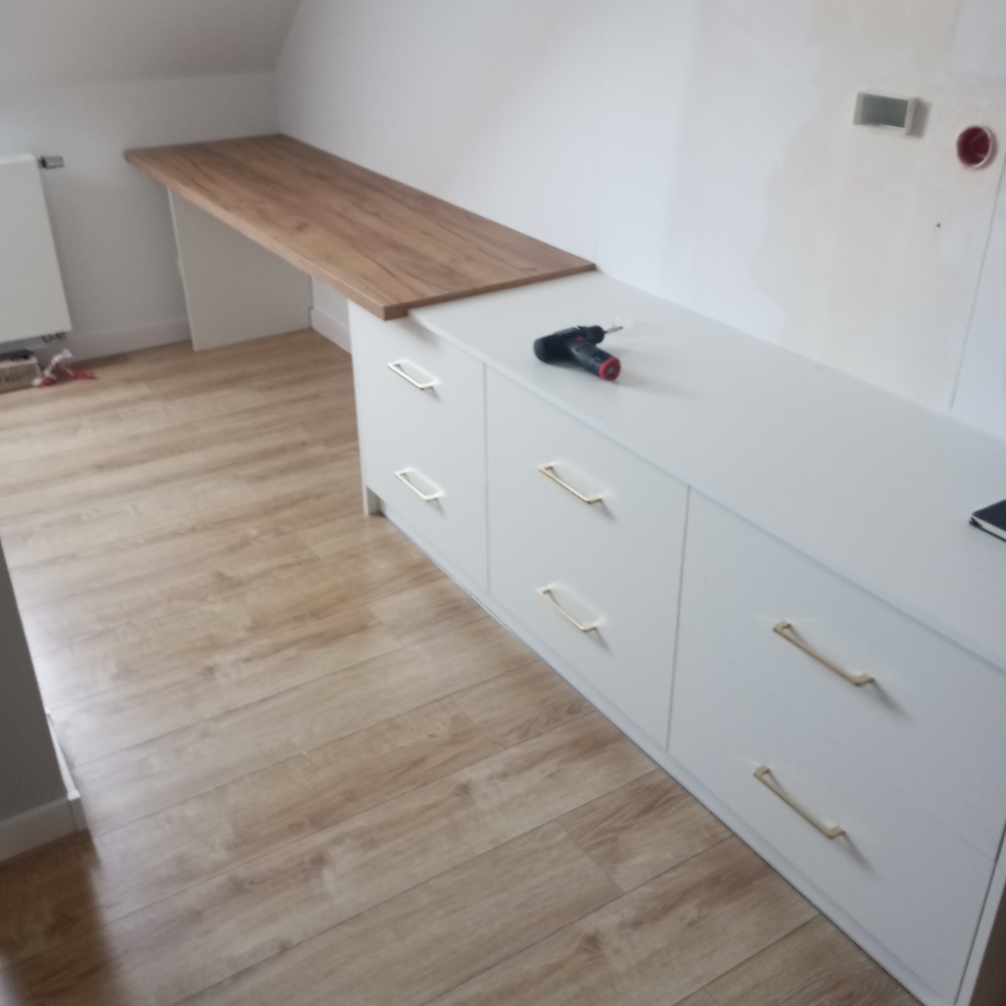 Biała komoda z szufladami i drewnianym blatem w skosie poddasza, złote uchwyty, wiertarka na blacie. Realizacja w jasnym wnętrzu.