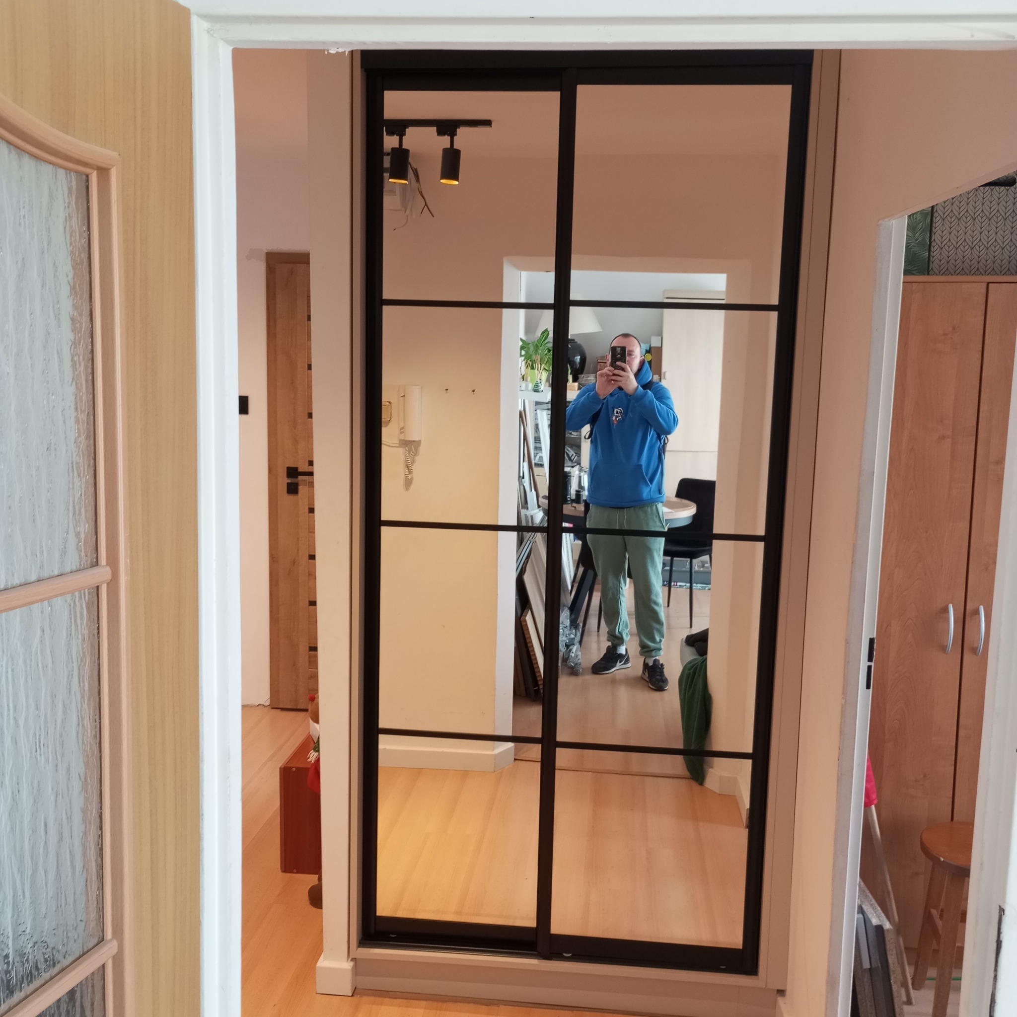 Nowoczesna szafa przesuwna z czarnymi szprosami, odbijająca wnętrze mieszkania i osobę robiącą zdjęcie. Widoczne drewniane podłogi i elementy wystroju.