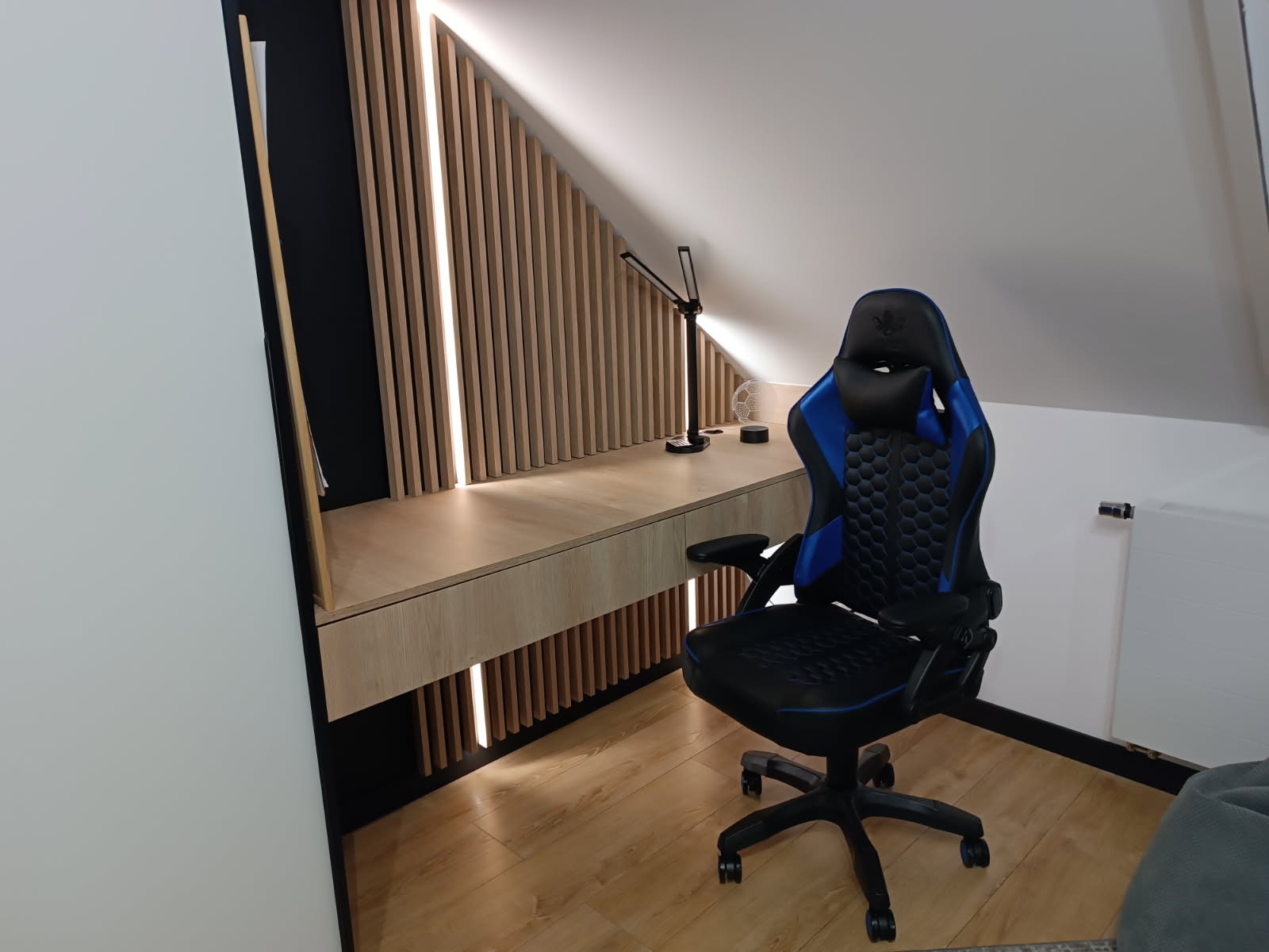 Biurko na poddaszu ze ścianką z lameli z podświetleniem LED, krzesło gamingowe. Jasne drewno, czarne akcenty. Nowoczesny design, oświetlenie punktowe.