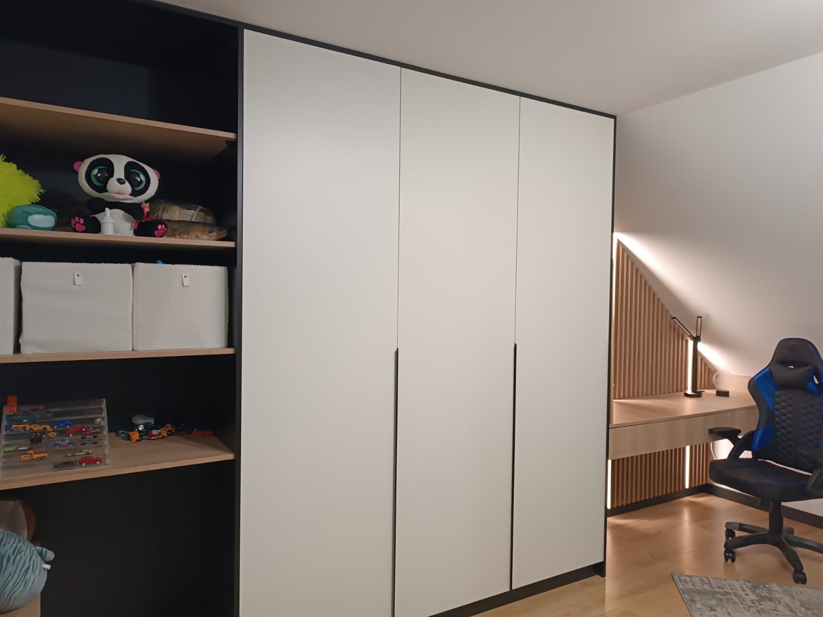 Biała szafa wnękowa z czarnymi akcentami w pokoju dziecięcym, widoczne półki z zabawkami i biurko z krzesłem gamingowym w tle.