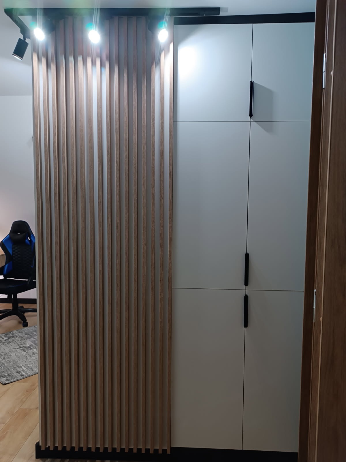 Biała szafa z czarnymi uchwytami i dekoracyjną ścianką z drewnianych listew w nowoczesnym wnętrzu. Oświetlenie punktowe na suficie.