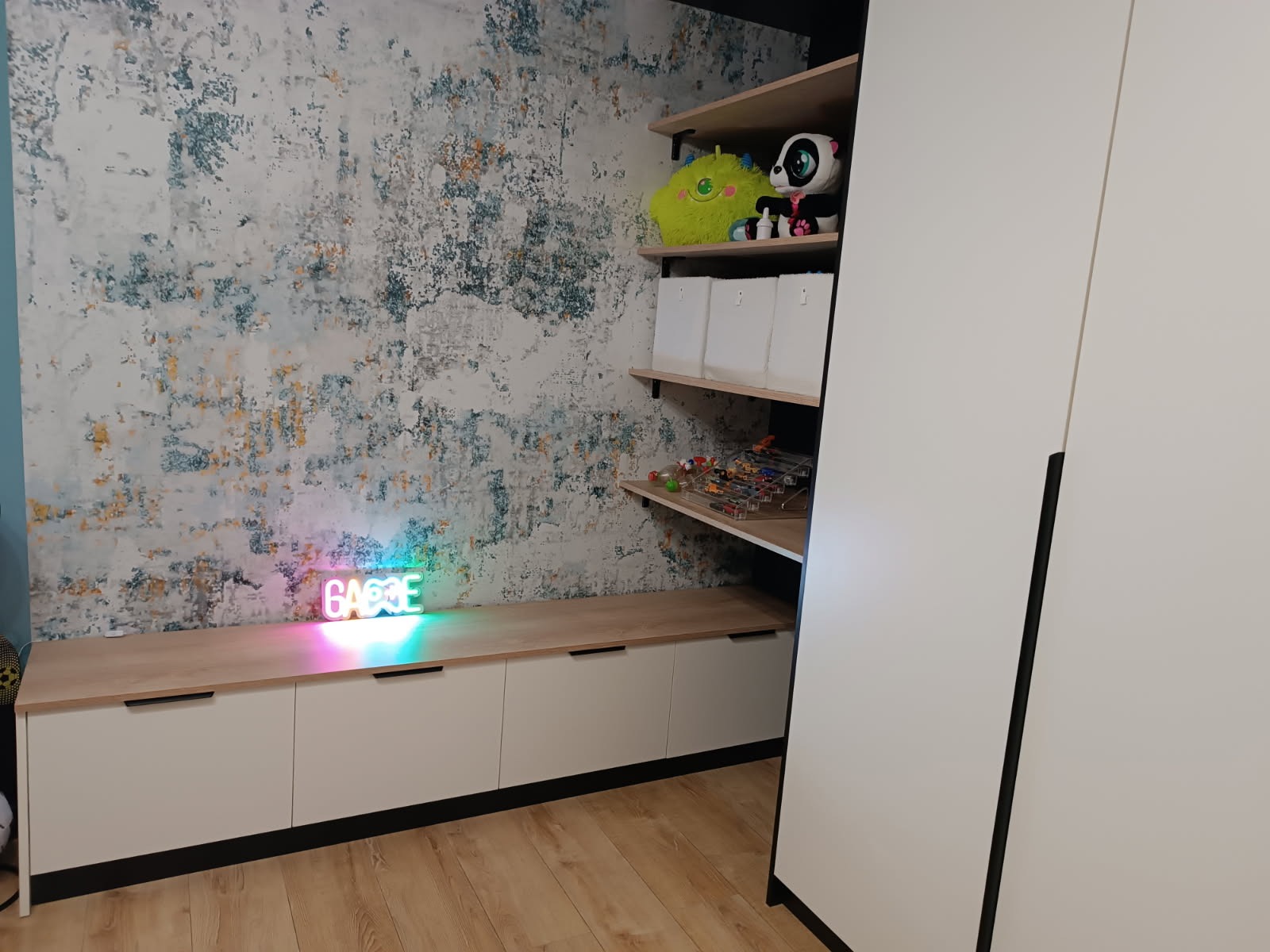 Biała szafa z półkami i szafkami pod telewizor, z czarnymi akcentami, na tle wzorzystej ściany i drewnianej podłogi. Na półkach zabawki i dekoracje.