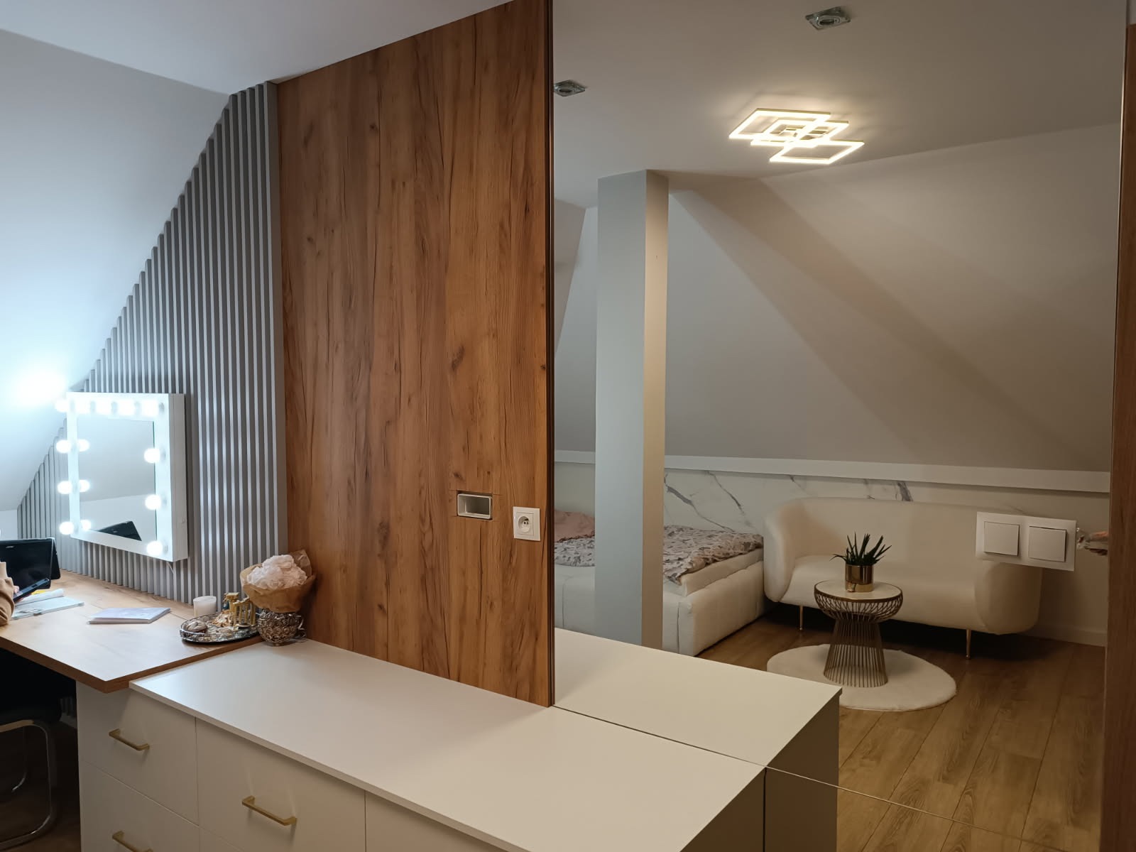 Stylowa garderoba w skosie z toaletką i lustrem do makijażu. Drewniana zabudowa z ukrytymi drzwiami, widok na pokój z sofą i marmurową ścianą w tle.