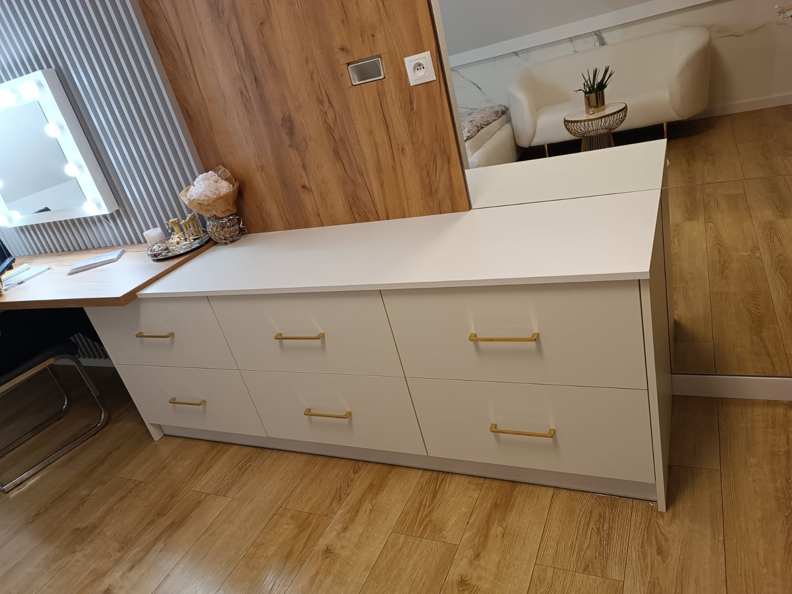 Biała komoda z sześcioma szufladami i złotymi uchwytami, część toaletki z lustrem i oświetleniem, drewniane panele na ścianie, odbicie sofy w lustrze.