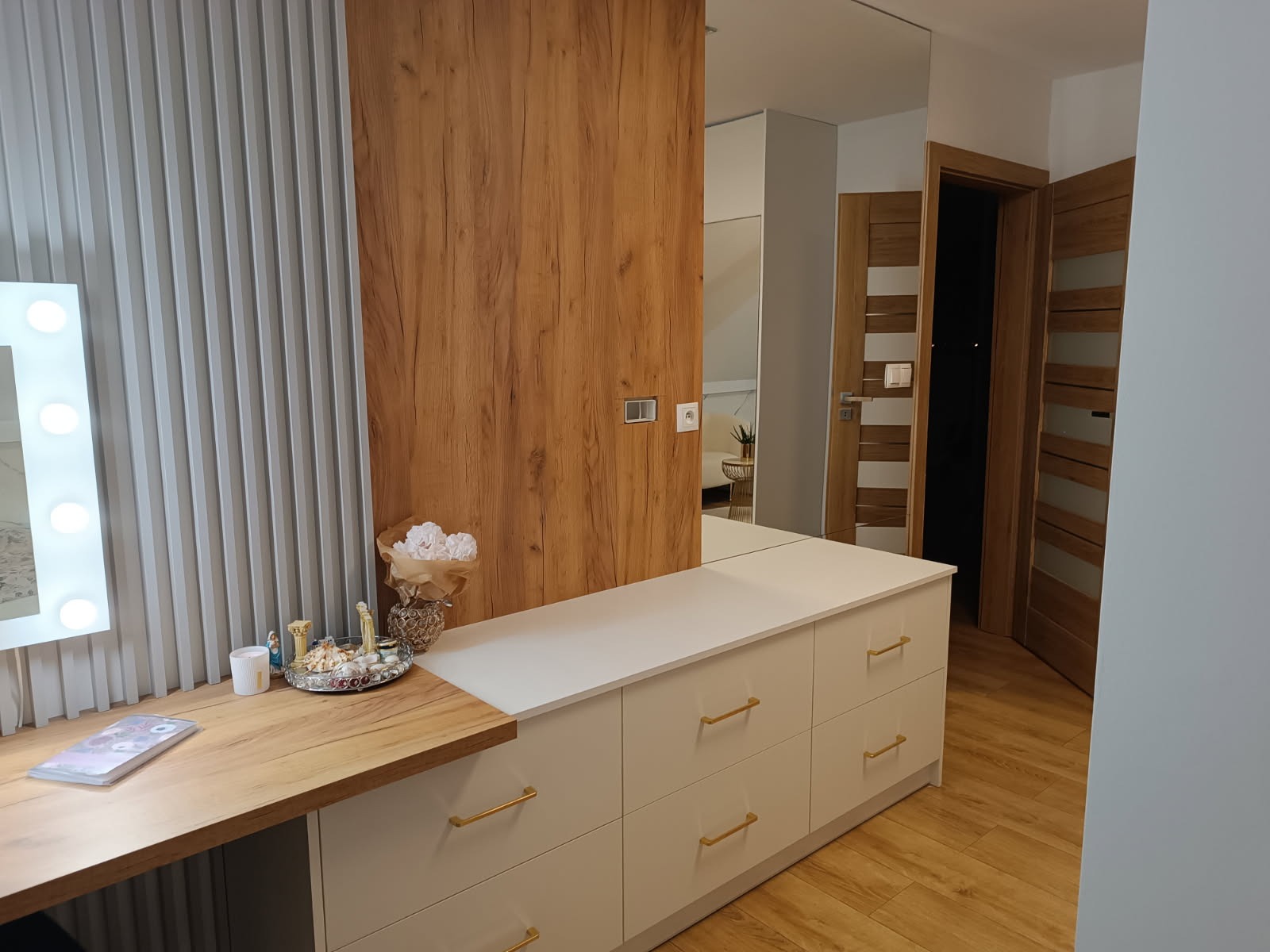 Biała komoda z szufladami i drewnianym blatem, na którym leżą drobne przedmioty. W tle lustro, drewniana ściana i drzwi. Całość w nowoczesnym stylu.