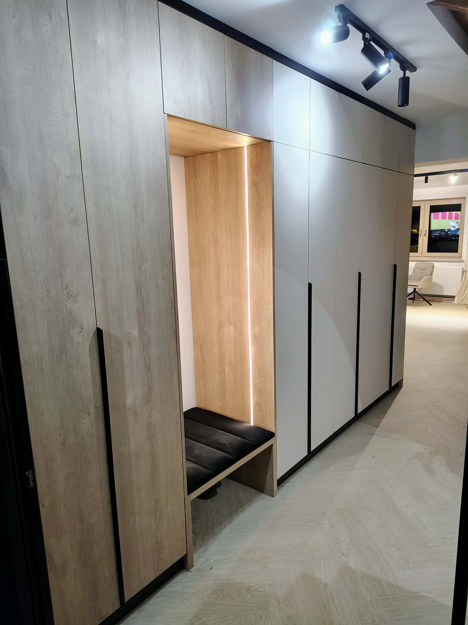 Szafa wnękowa z siedziskiem i oświetleniem LED. Fronty w odcieniach szarości i beżu, czarne uchwyty. Widoczne oświetlenie sufitowe i podłoga w jodełkę.