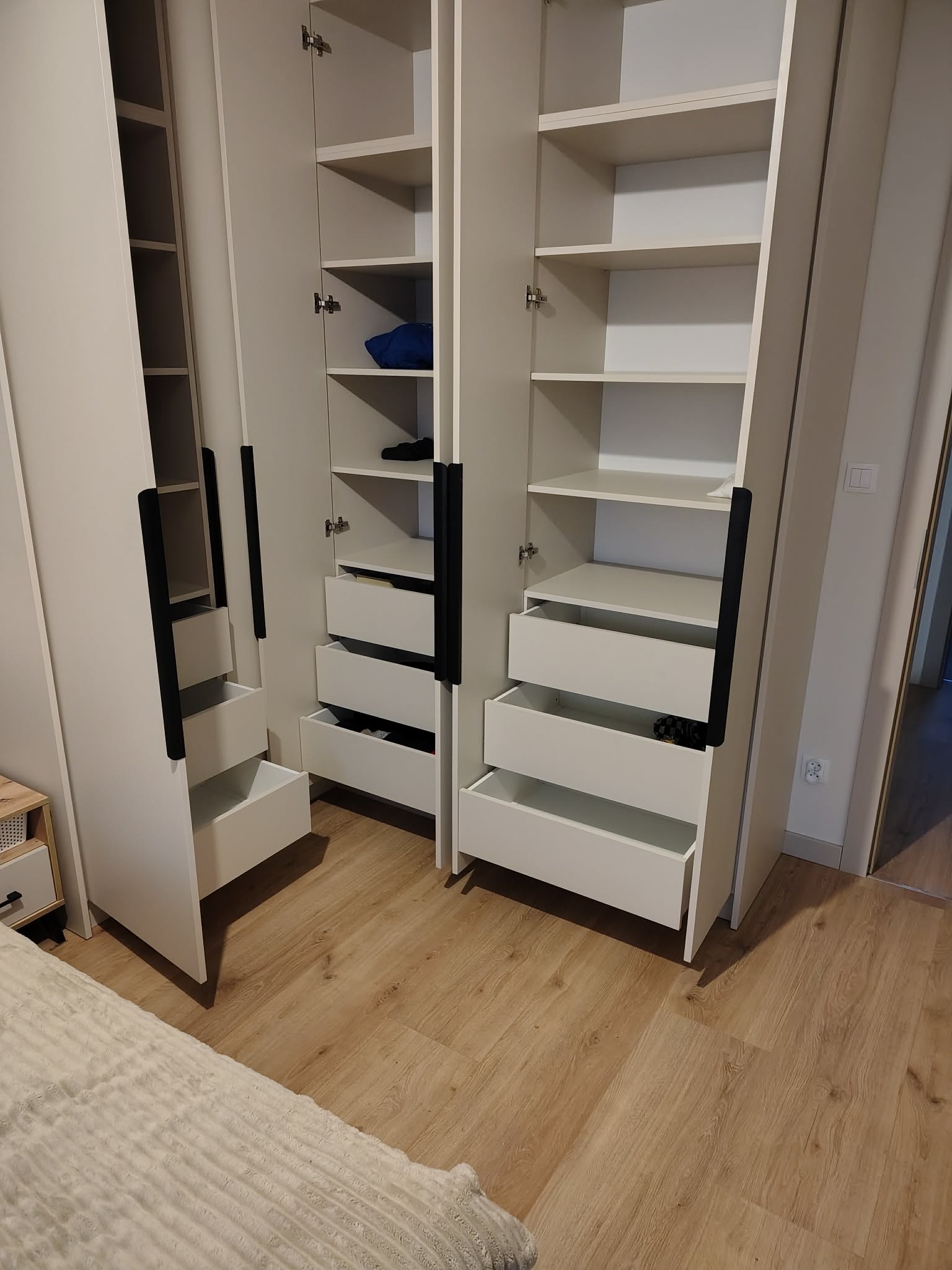 Biała szafa wnękowa z czarnymi uchwytami, otwarte drzwi i szuflady ukazują półki i zawartość. Jasne drewno podłogi, fragment łóżka w kadrze.