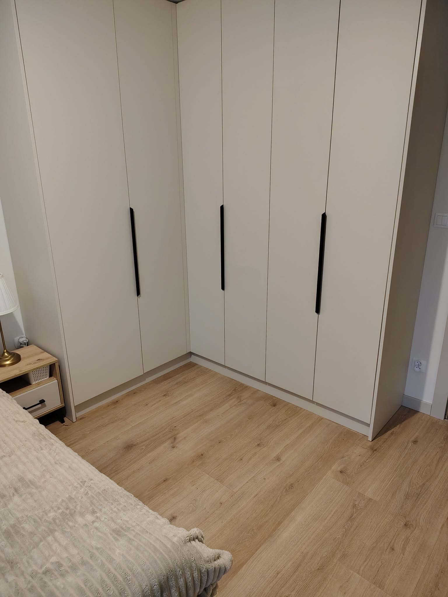 Jasna szafa wnękowa z czarnymi uchwytami, dopasowana do rogu pokoju, z widoczną częścią łóżka i szafki nocnej. Podłoga z paneli w odcieniach drewna.