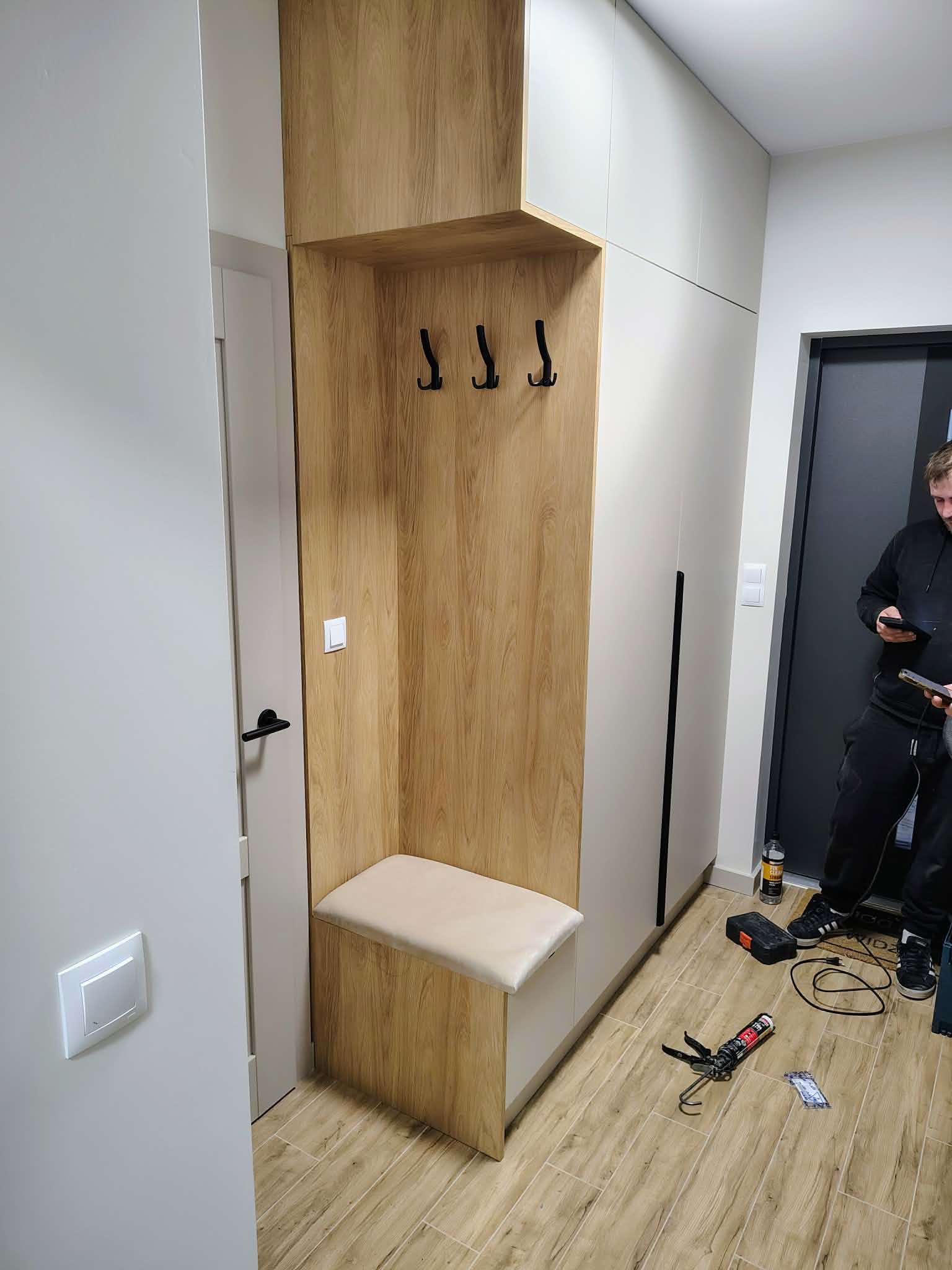 Funkcjonalna szafa w przedpokoju z siedziskiem i wieszakami, wykończona w drewnie i bieli. Widoczne narzędzia montażowe na podłodze.