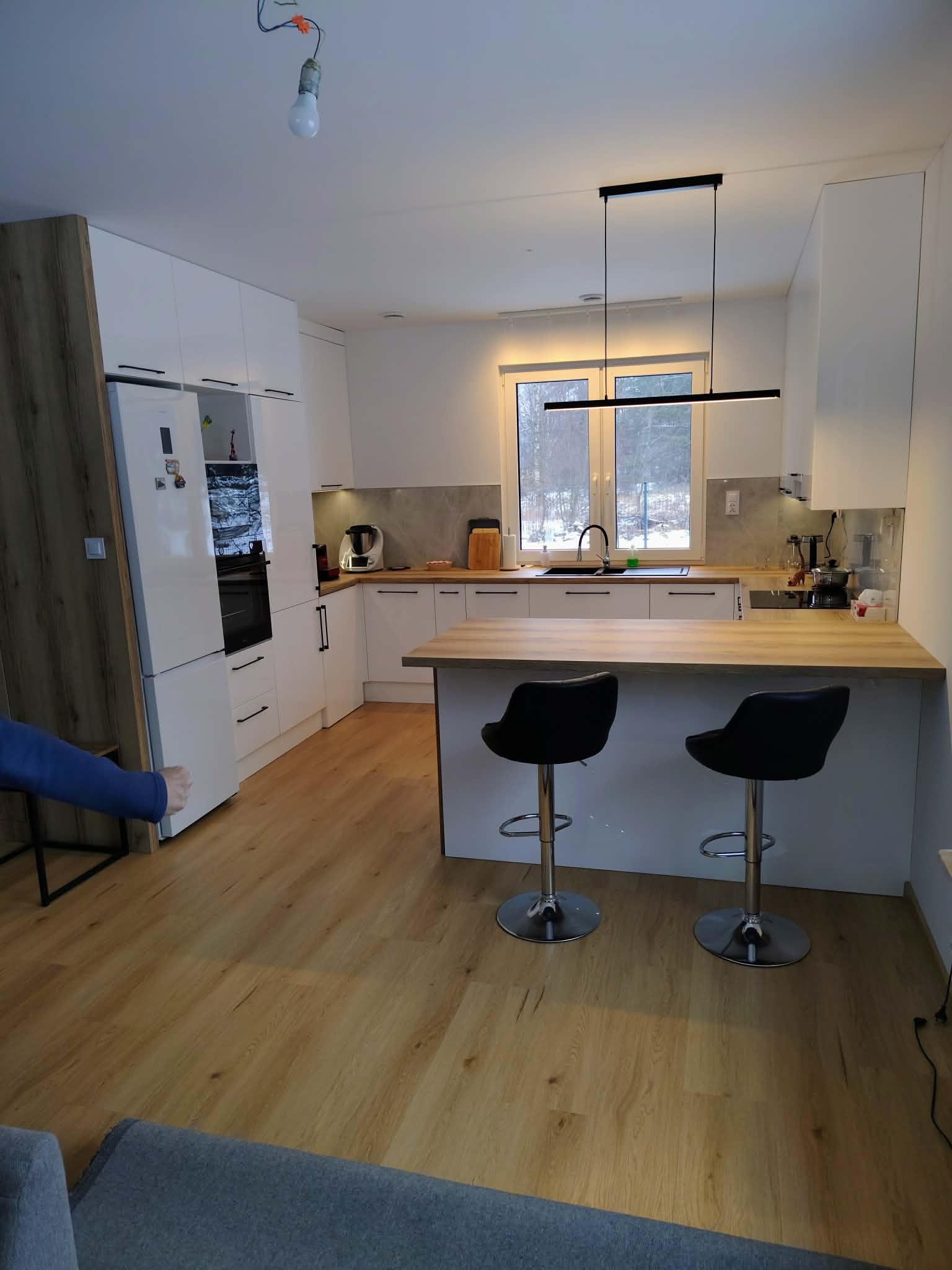 Jasna kuchnia z wyspą i dwoma hokerami. Białe fronty szafek, drewniany blat. Nad wyspą nowoczesna, czarna lampa. Widoczne okno z widokiem na zewnątrz.