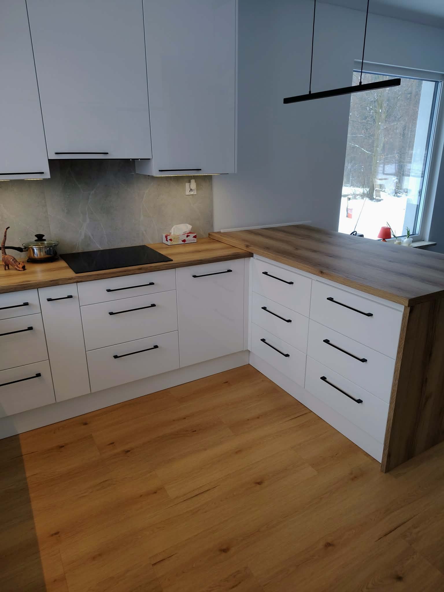 Jasna kuchnia z drewnianym blatem i czarnymi uchwytami, widok na okno z zimowym krajobrazem. Minimalistyczny design, nowoczesne oświetlenie.