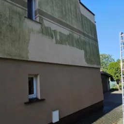 Fragment elewacji budynku z widocznymi obszarami po częściowym usunięciu zabrudzeń, zarys drabiny oparty o ścianę oraz fragment chodnika z kostki brukowej.