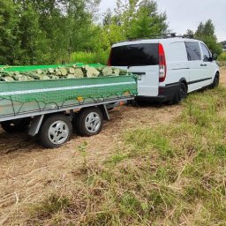 Instaltec - Przyczepa wypełniona pociętymi kawałkami drewna, zabezpieczona zieloną siatką, ciągnięta przez biały samochód dostawczy na trawiastym terenie.