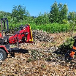 Instaltec - Pracownik w ochronnikach słuchu obsługuje rozdrabniacz gałęzi, z którego wylatują wióry, w tle zielona roślinność.