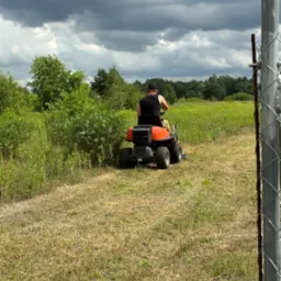 Operator na pomarańczowym traktorku kosi wysoką trawę na łące w pochmurny dzień, widok zza ogrodzenia z siatki.