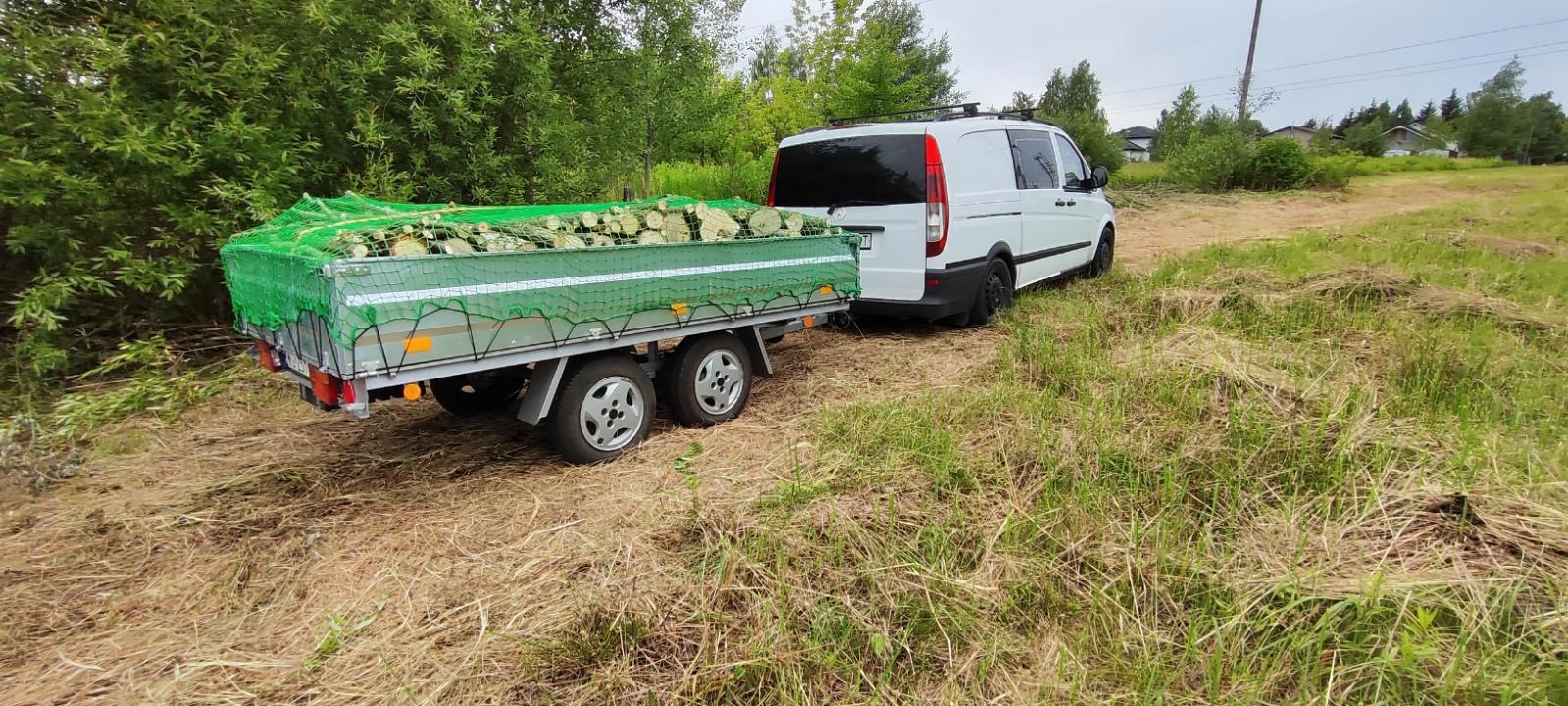 Przyczepa wypełniona pociętymi kawałkami drewna, zabezpieczona zieloną siatką, ciągnięta przez biały samochód dostawczy na trawiastym terenie.
