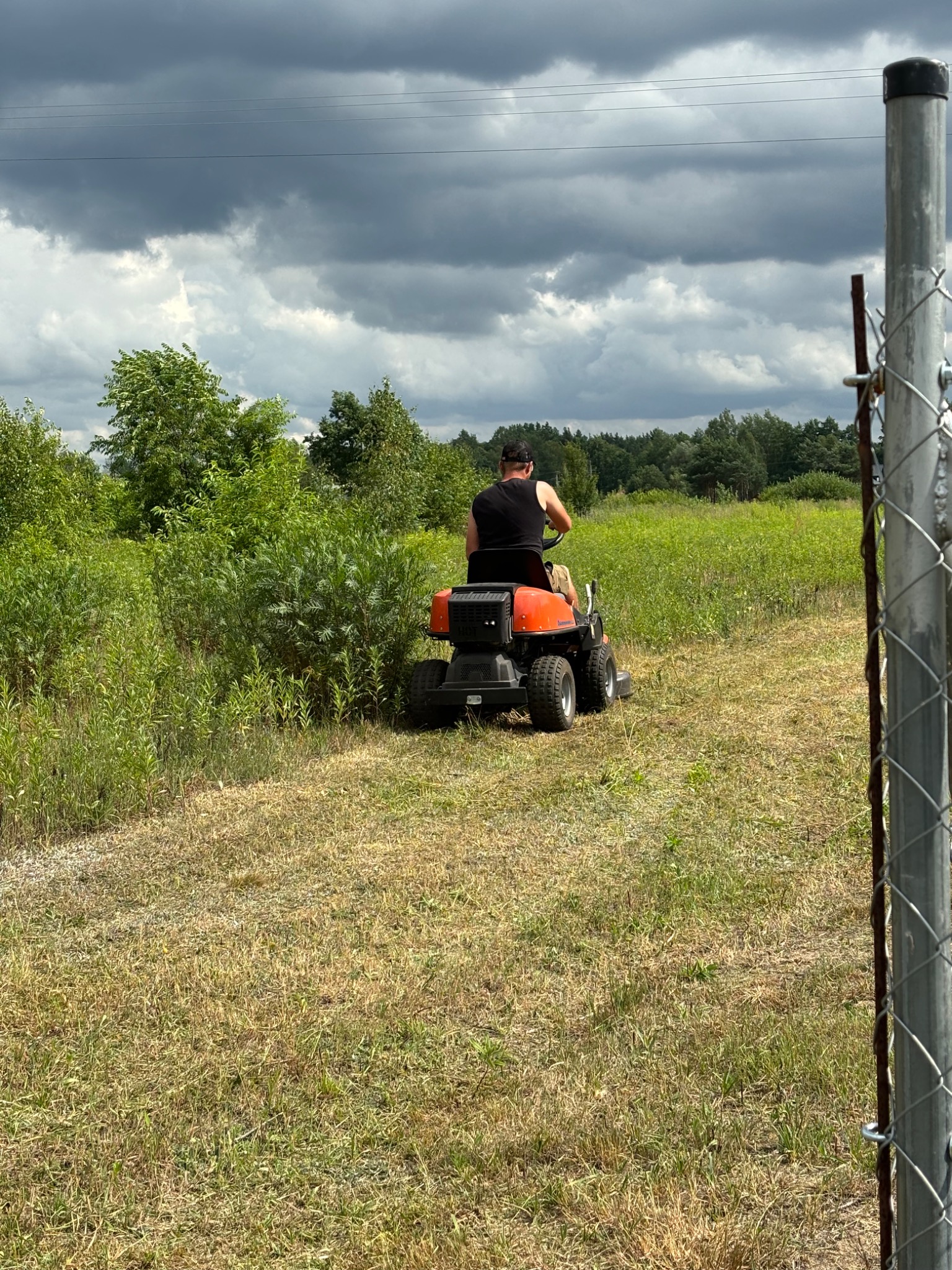 Operator na pomarańczowym traktorku kosi wysoką trawę na łące w pochmurny dzień, widok zza ogrodzenia z siatki.