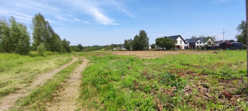 Rozległy teren z wykoszoną trawą, przygotowany pod budowę, z widoczną polną drogą i zabudowaniami w tle.