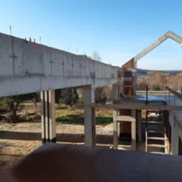 Surowa konstrukcja budynku w trakcie budowy z widocznym teodolitem na tle krajobrazu.