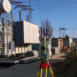 Pomiar geodezyjny z użyciem teodolitu na statywie przed transformatorem wysokiego napięcia na tle słupów energetycznych i komina przemysłowego, w tle pracujący robotnicy.