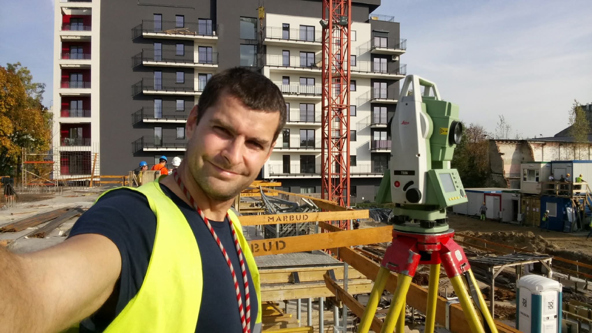 Uśmiechnięty mężczyzna w kamizelce odblaskowej robi selfie z niwelatorem na budowie. W tle budynek mieszkalny i dźwig.