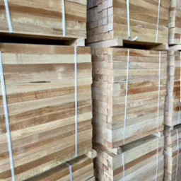 Stosy tarcicy budowlanej z jasnego drewna, zabezpieczone białymi taśmami, ułożone w magazynie.