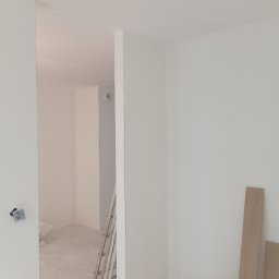 Jakub Jabłoński - Wnętrze w trakcie remontu: białe ściany po szpachlowaniu, drabina oparta o ścianę, worek z zaprawą i deski podłogowe w rogu, widoczny gniazdko elektryczne.