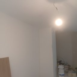 Jakub Jabłoński - Wnętrze w trakcie remontu z widocznymi świeżo wyszpachlowanymi ścianami, wiaderkami z farbą i odsłoniętym przewodem elektrycznym zwisającym z sufitu.