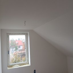 Jakub Jabłoński - Poddasze po pracach wykończeniowych: świeżo pomalowane ściany w odcieniach bieli, okno z widokiem na zabudowę jednorodzinną, zabezpieczone folią parapety, widoczne gniazdko elektryczne...