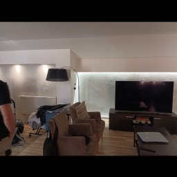 Jakub Jabłoński - Salon z szarą ścianą, oświetleniem LED, brązowymi fotelami, telewizorem na szafce RTV i podłogą z paneli. W tle widoczny grzejnik i rzeczy osobiste, sugerujące trwające prace.